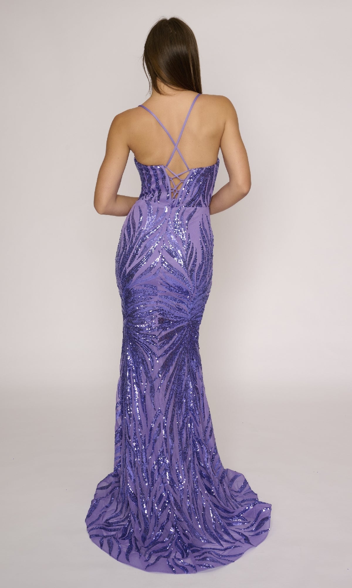 A woman models the Nina Canacci 4433 long formal dress—a floor-length, sleeveless purple sequin prom gown with a fitted silhouette and lace-up crisscross open back—shown from behind against a plain background.